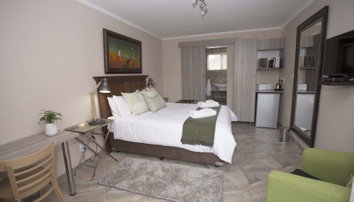A guest room with a queen bed, a desk under the window and a kitchenette along the far wall.
